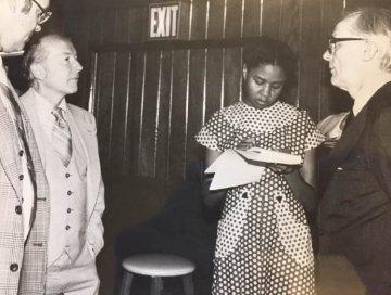Black and white photo of 4 people talking, a black girl dressed in a pocadot dress is taking notes on a notebook