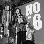 A black and white photo of Sally talking into a microphone in front of a backdrop that says No on 6