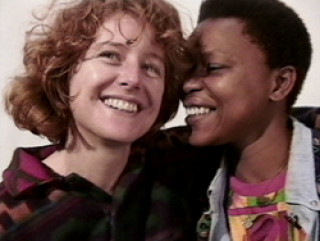 A red-haired caucasian woman and an African woman embrace while smiling and laughing.