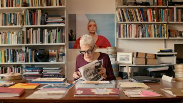 Florrie Burke sits at a table with bookshelves in background and looks through her former partner Barbara Hammer's artwork.
