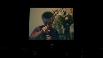 A self-portrait of and by Barbara Hammer with her pointing at the camera and flowers in the background.