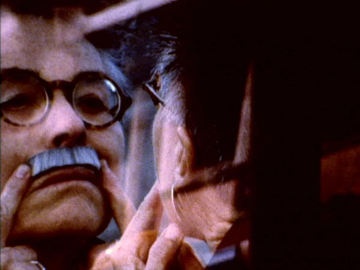 Barbara Hammer puts on a mustache as she looks in a mirror.