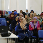 A group of hijabi women sit on blue chairs, observing something happening in front of them, some of them laughing, some keeping a straight face